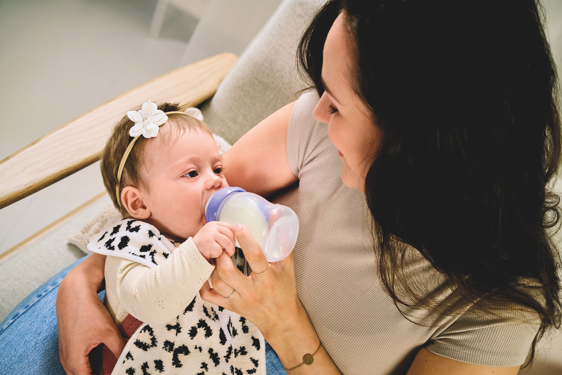 Adult female holding infant with bottle, infant wears patterned bib with floral headband.