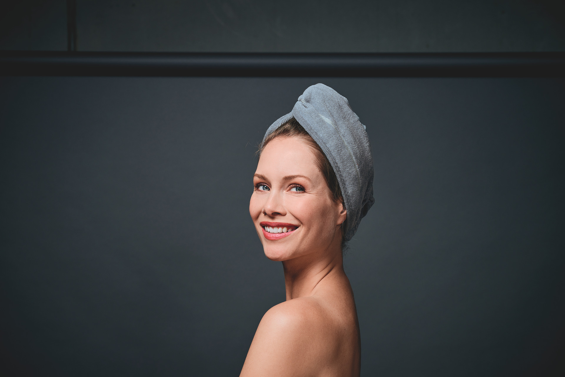 Adult woman with towel wrapped around her head, smiling against a dark background.