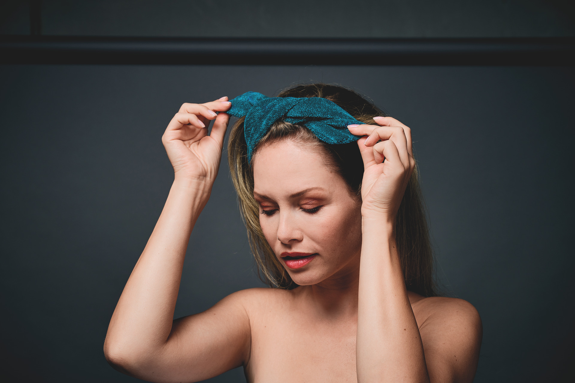 Adult woman with long hair adjusts a teal fabric headband. Plain backdrop. Focus on accessory.