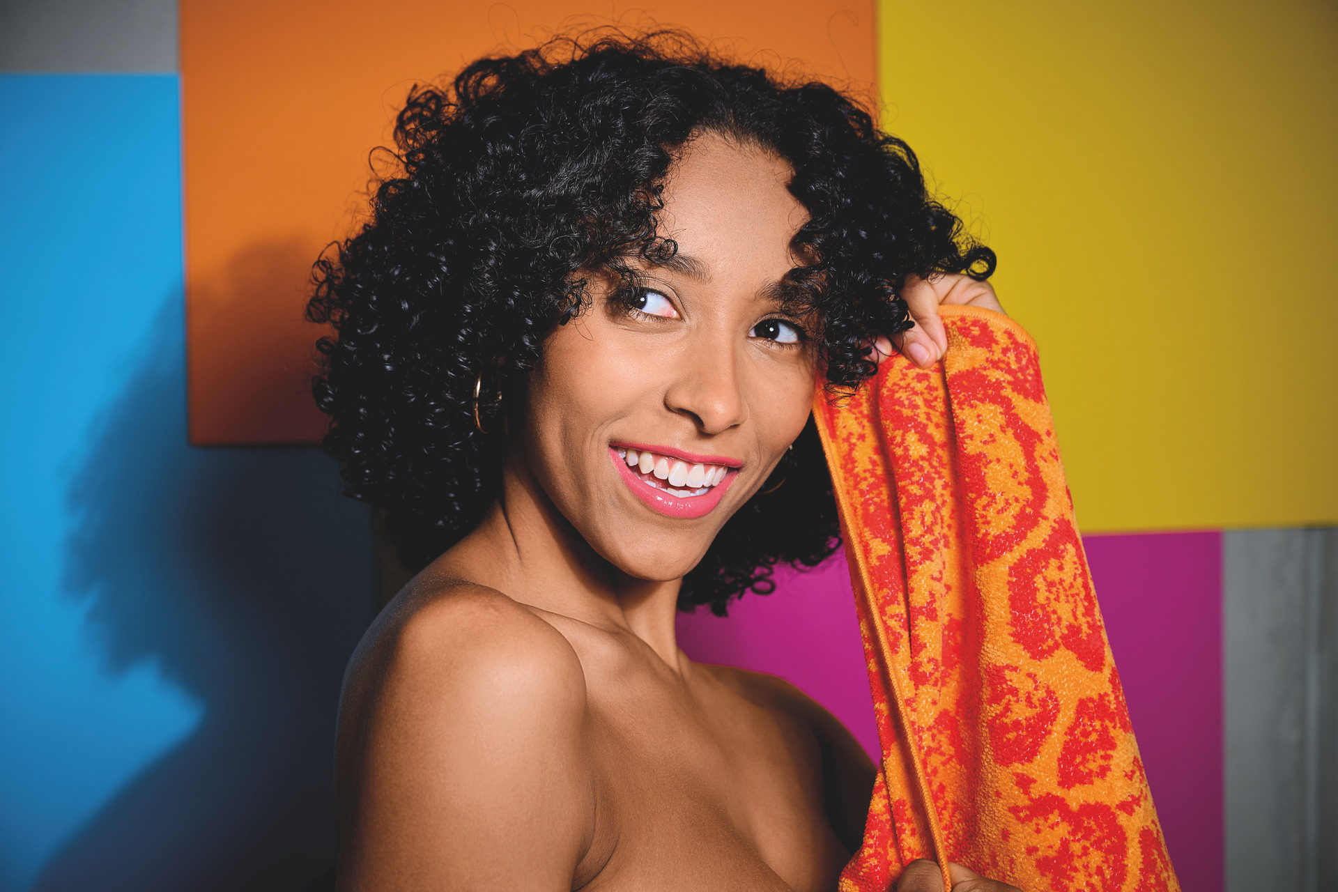 Adult female with curly hair holding orange-red textured fabric, standing against a colourful background.
