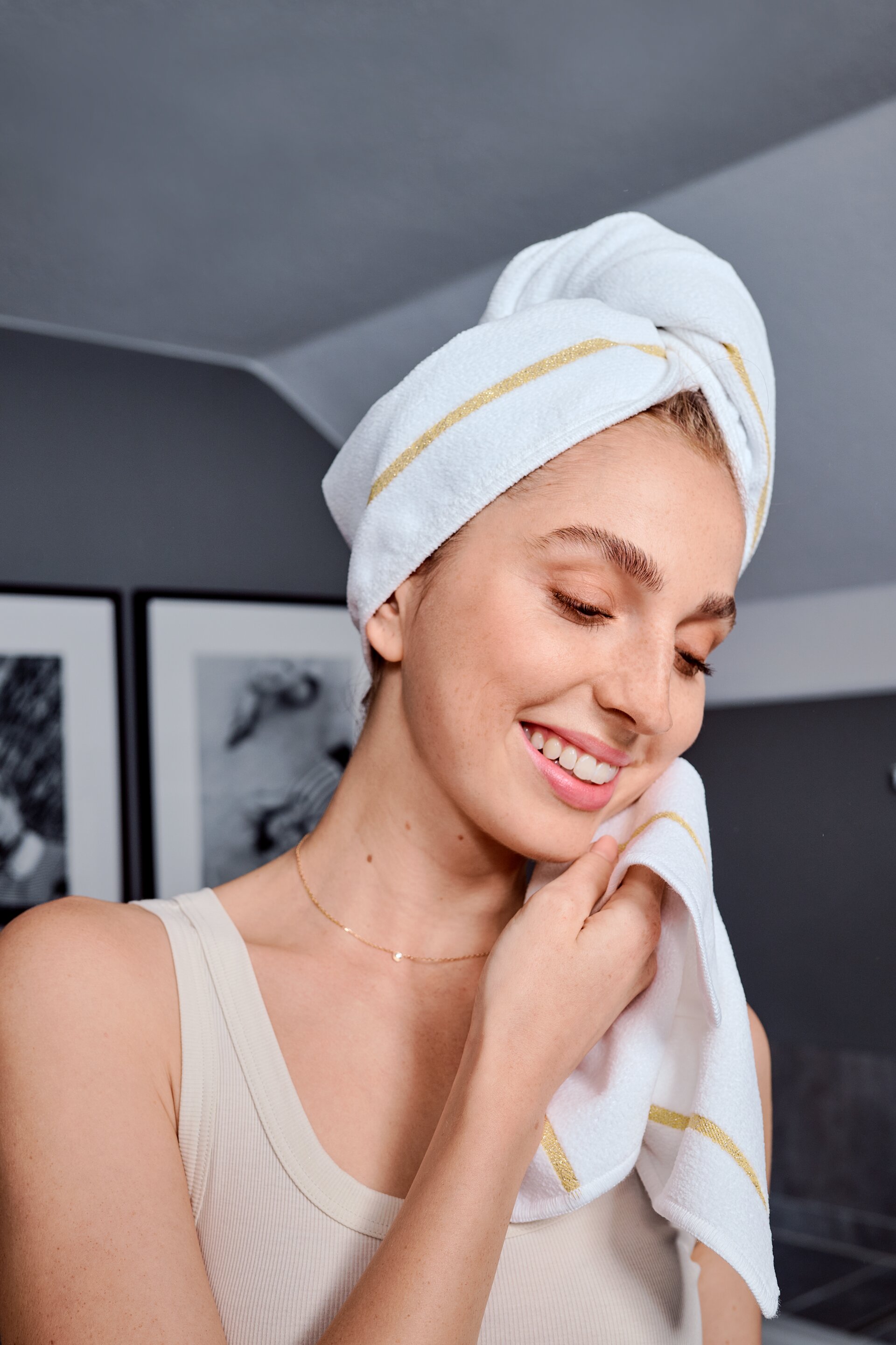 Adult woman with white towel, golden stripes; smiling in neutral-toned room. Short hair wrapped in towel.