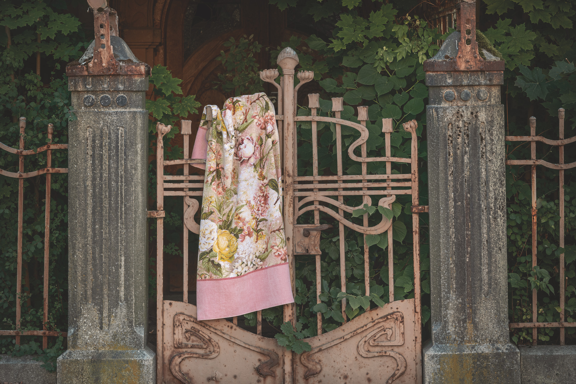 Ornate textile draped on ornate gate; floral pattern with pink border, set against lush greenery. No models visible.