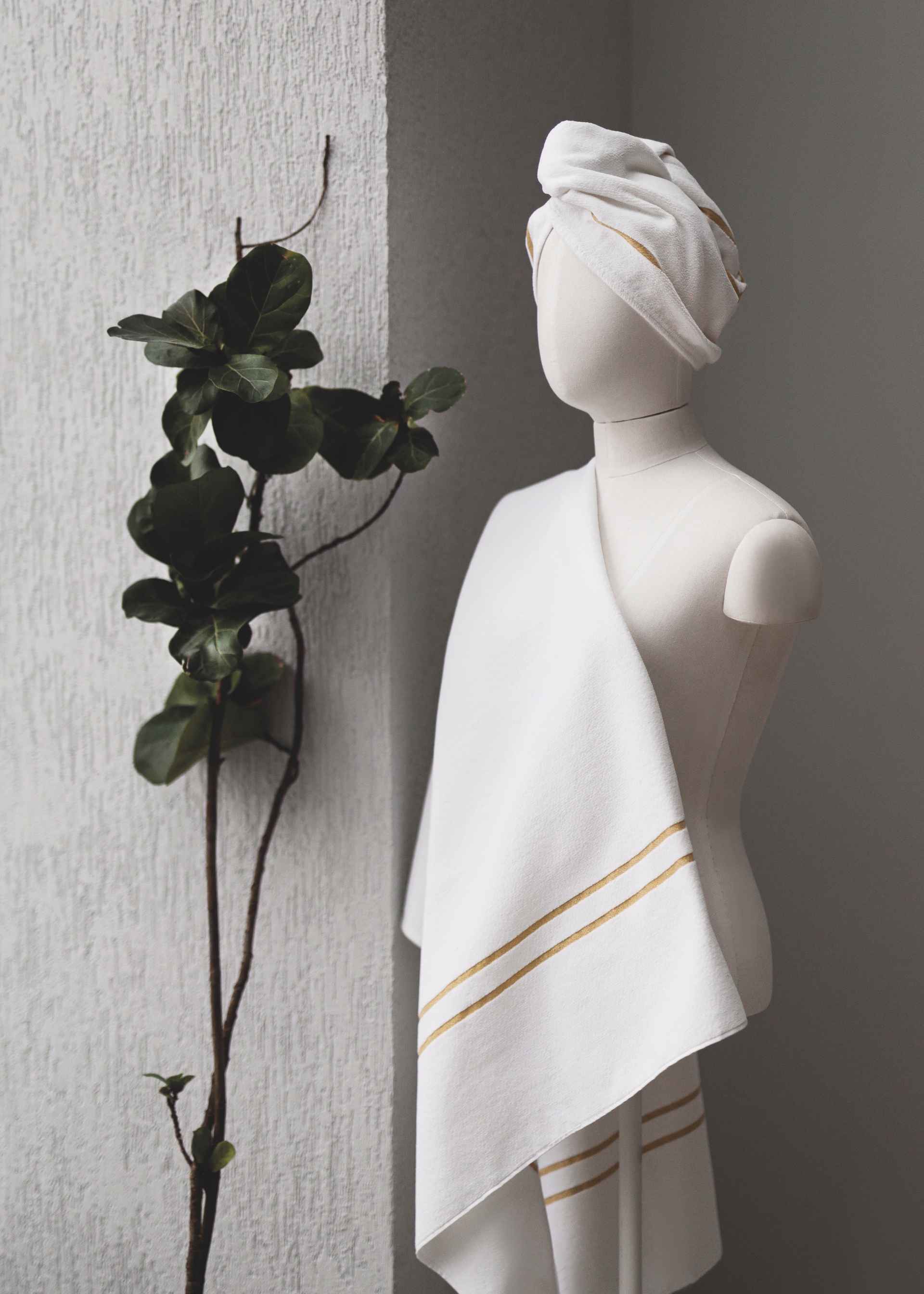 Adult mannequin with white, gold-striped fabric and head wrap, beside green plant on a textured wall.