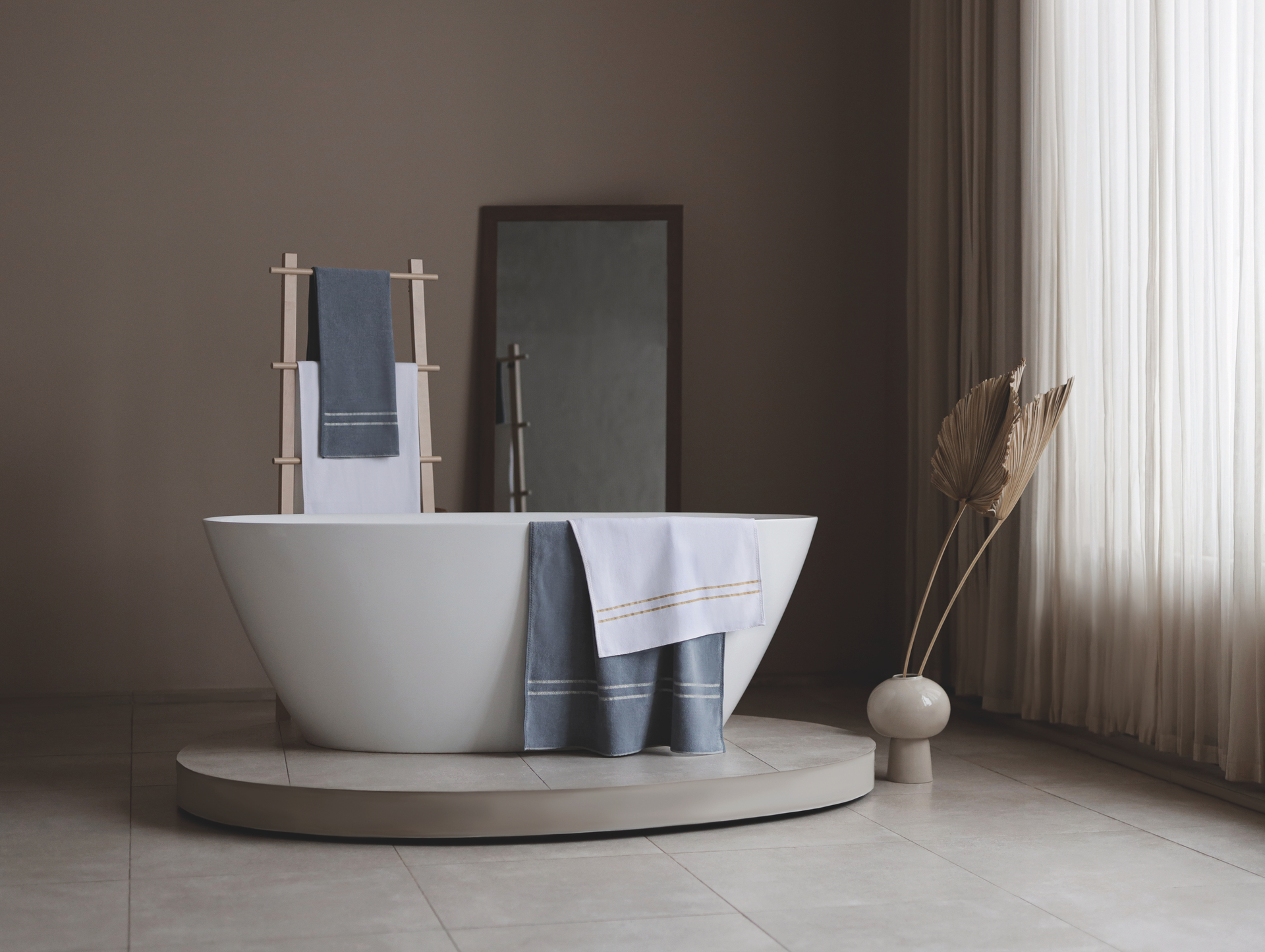 Modern bathroom with a white freestanding tub, grey and white towels hanging, and a minimalistic brown decor.