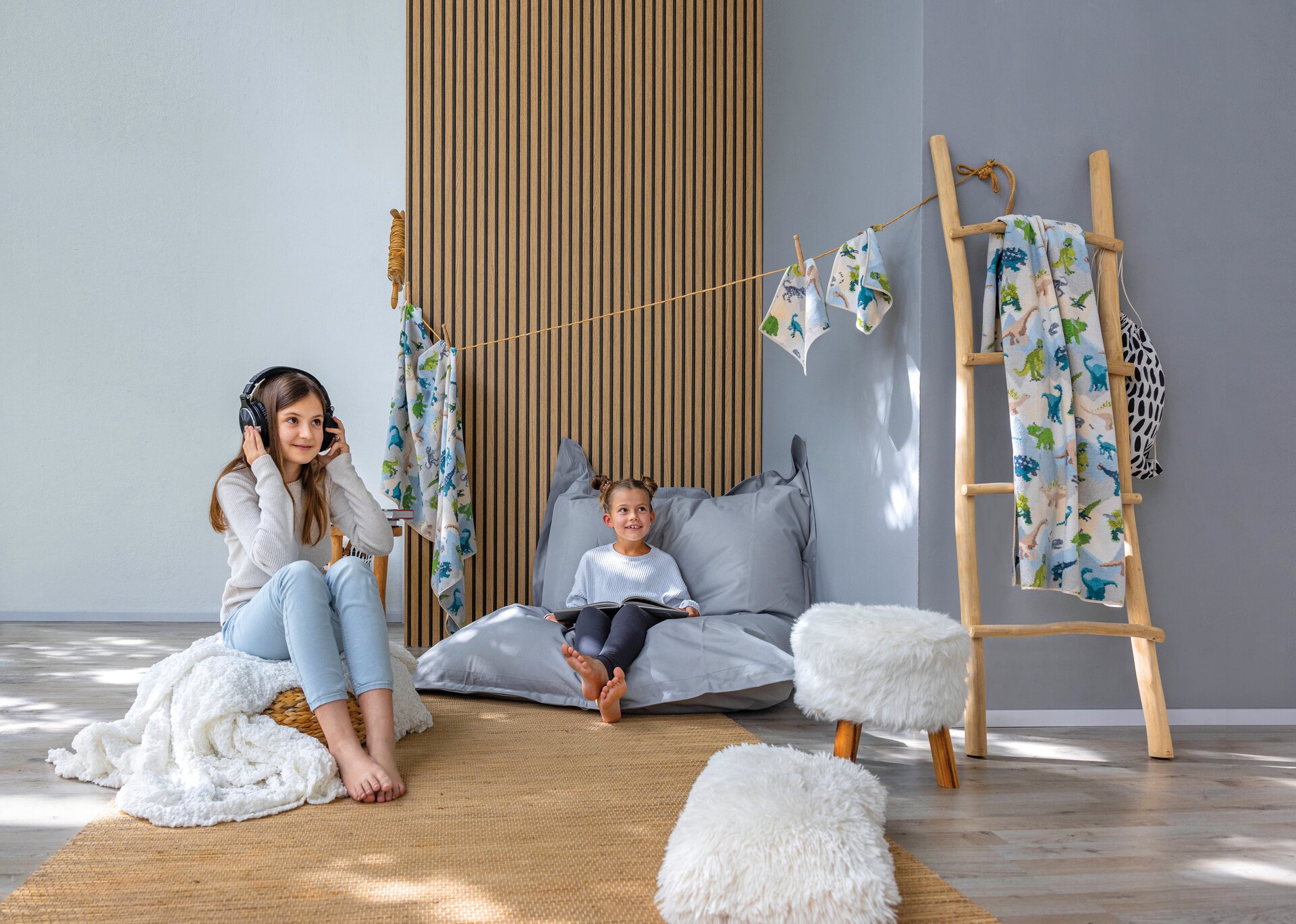 Two children in a bright room, with nature-patterned textiles hanging; one listening to music, the other on a beanbag.