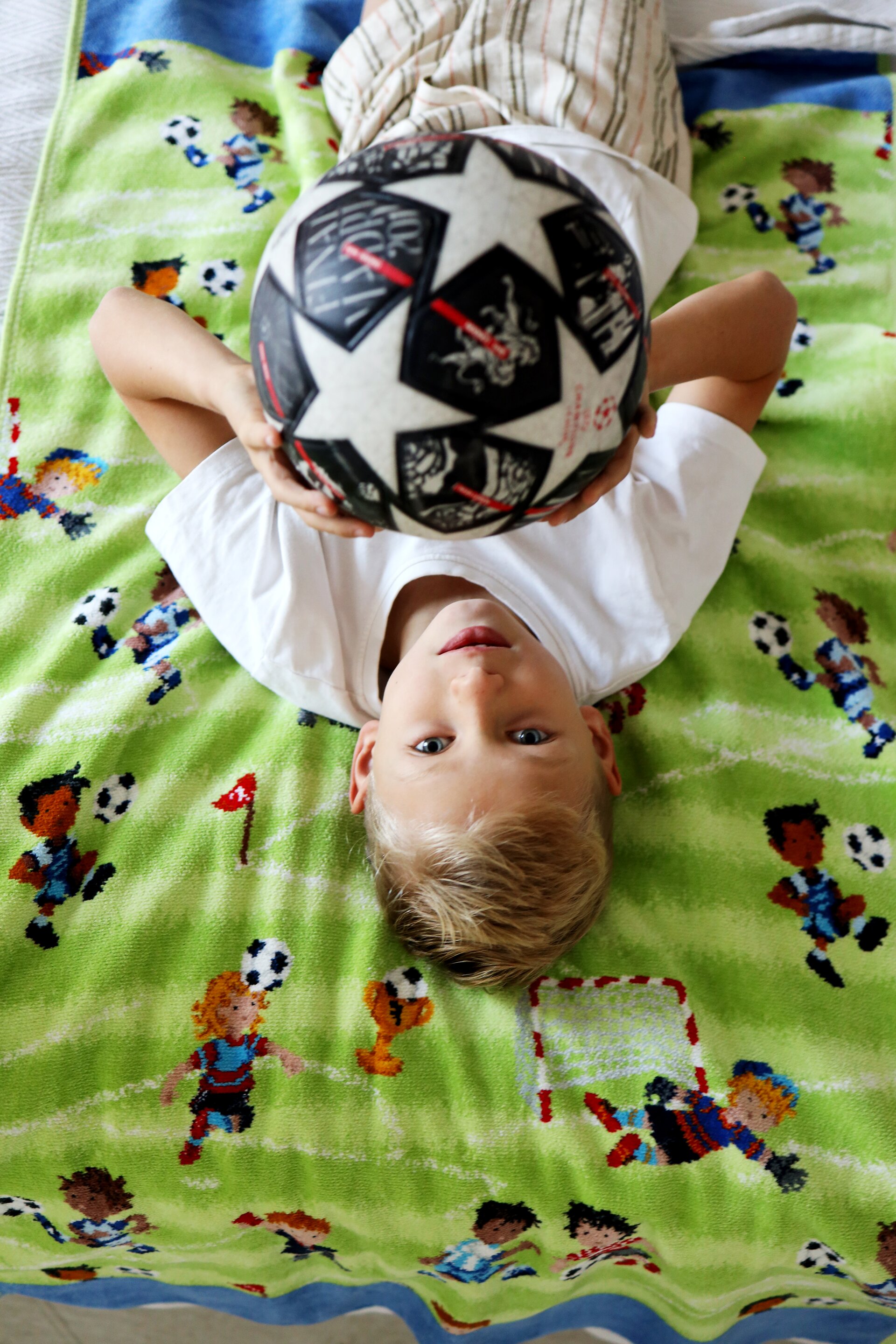 Young boy holding a football, lying on a vibrant textile with playful children and football pattern.
