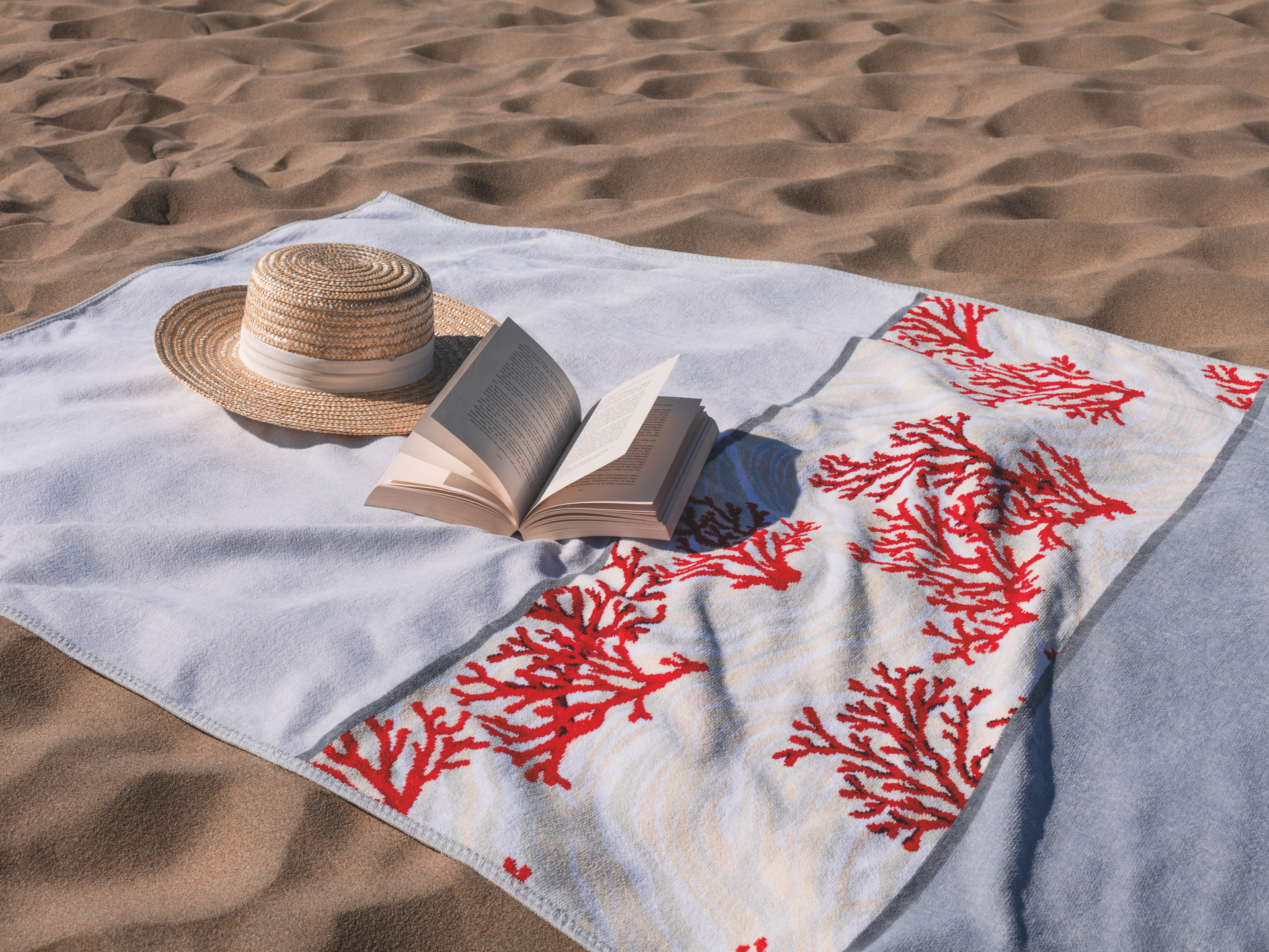 Towel with red coral pattern on sand, accompanied by a straw hat and an open book; elegant beach setting.
