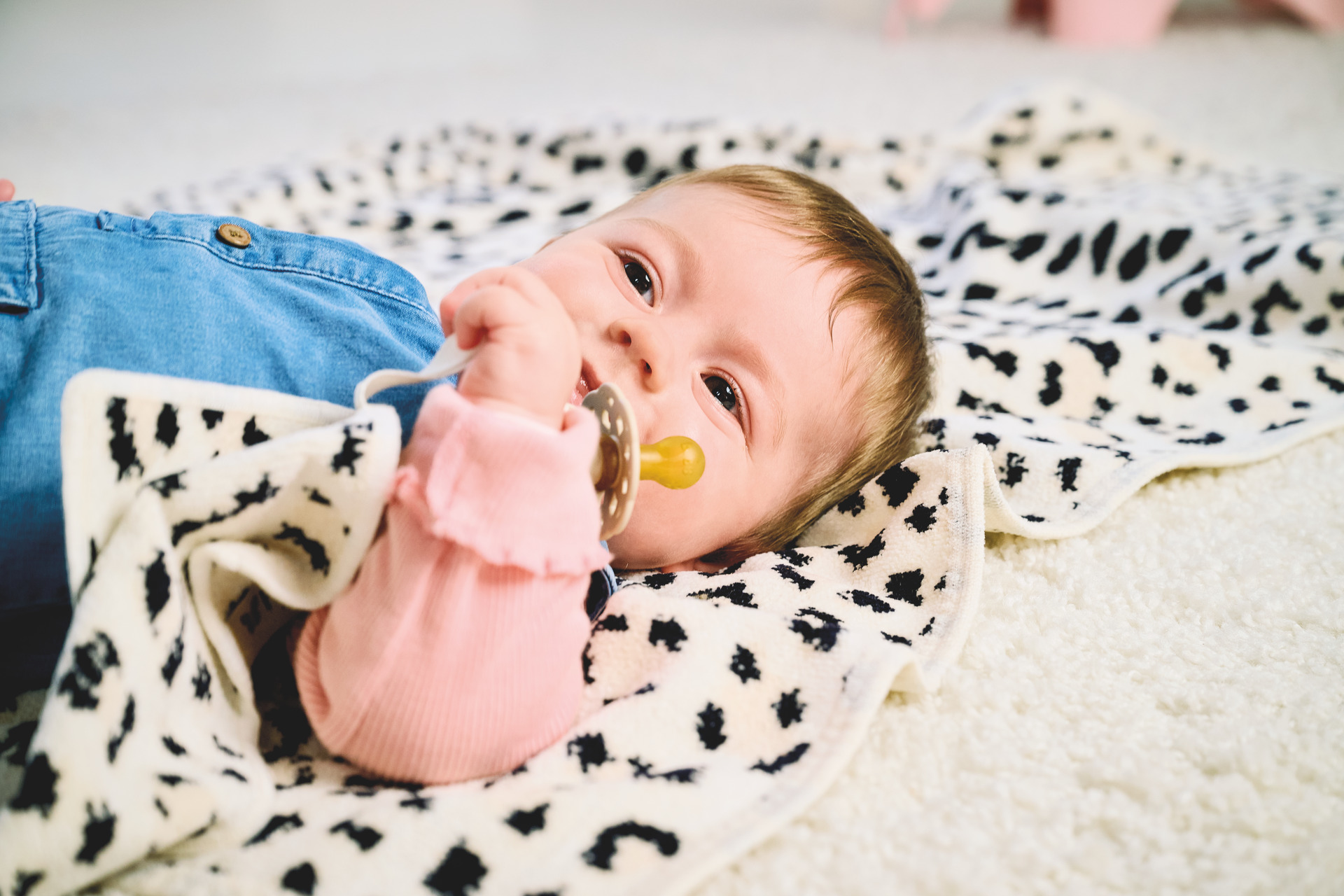 Baby lying on a soft white fabric with black abstract patterns; wearing a pink sleeve and denim overalls.