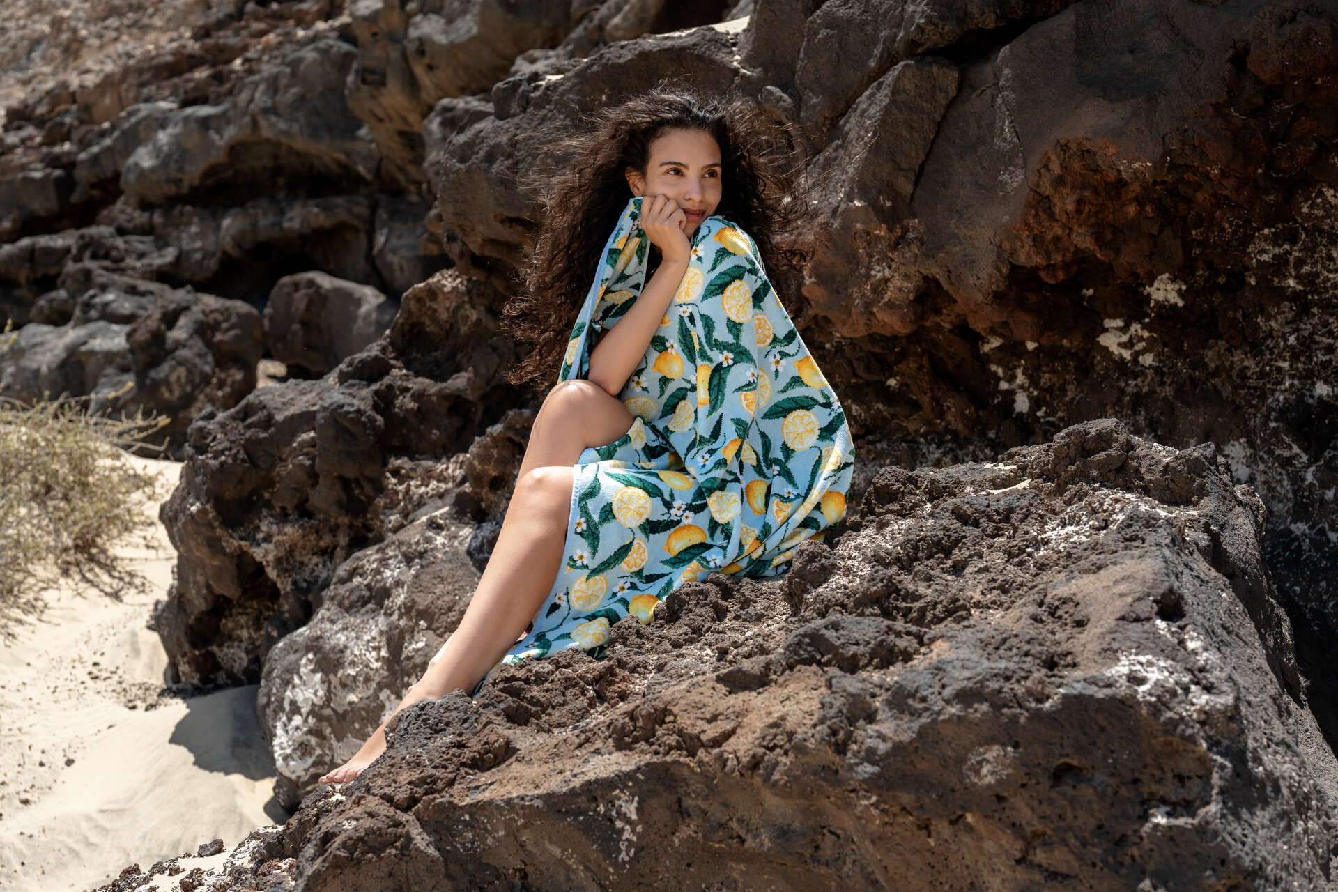 A woman sitting on rocks at the beach, wrapped in a colorful shawl with a lemon pattern.